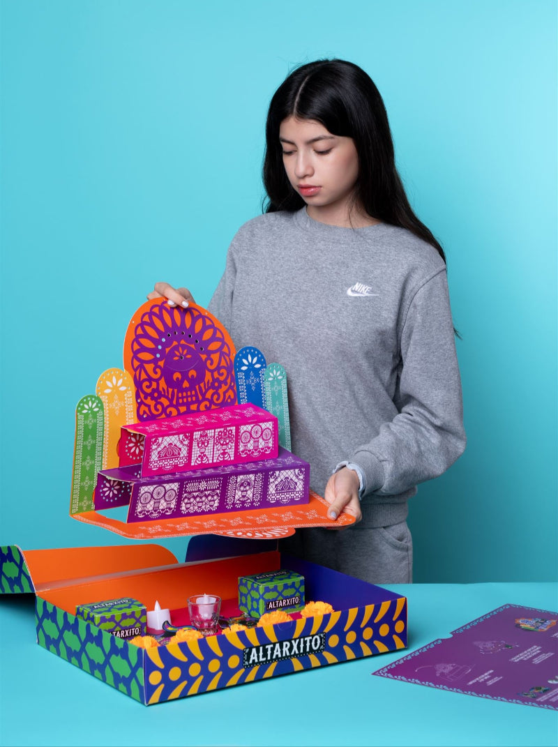 a girl with dark hair holds a colorful altar above a box labeled altarxito with candles and flowers
